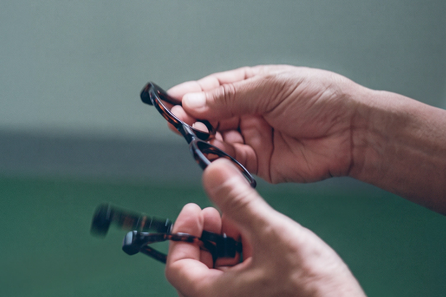 Two hands holding a pair of dark tortoiseshell-pattern eyeglass frames, one arm of the frames blurred by motion, against a soft green-grey background